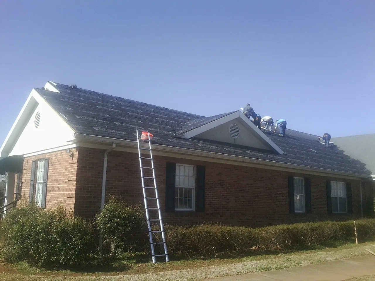 Crew tearing shingles off a brick ranch roof with a ladder set — Metal Roof Repair work by our crew in Duncombe