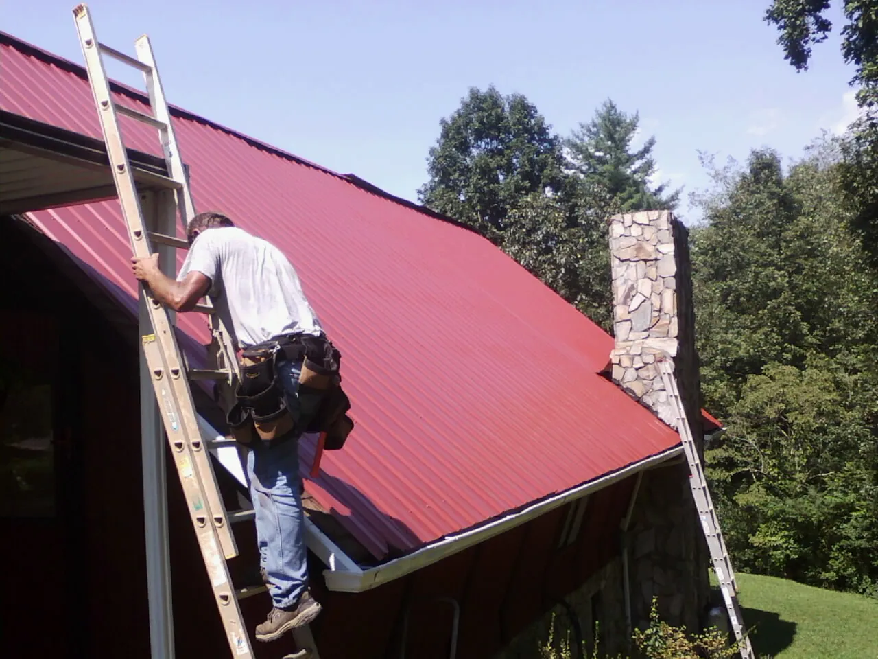 Roofer on a ladder beside a red metal roof and stone chimney — Soffit & Fascia Repair by Roofing Brothers in Duncombe